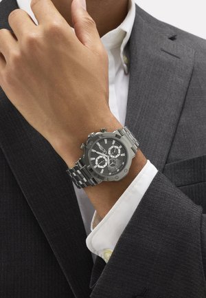 Man in donker pak die zijn kraag aanpast, polshorloge met zwarte wijzerplaat en zilveren metalen band aan zijn linkerpols.