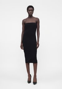 Femme portant une robe midi noire sans bretelles, ajustée, et des escarpins noirs à bouts pointus, debout sur un fond blanc uni.