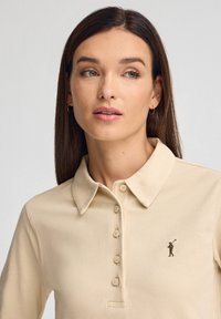 Polo beige con cuello con solapa, presenta una fila de botones y un pequeño logo bordado de un golfista en el pecho.