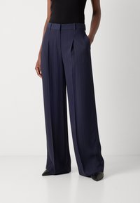 Pantalons rayés bleu marine avec un design à jambes larges, des plis à l'avant et une finition lisse. Portés avec un haut noir et des chaussures à bout pointu.