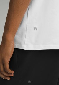 Hvid bomulds t-shirt med korte ærmer og lige kant, som har et lille logo på den nederste side, sat sammen med sorte bukser.