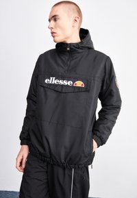 Giacca a vento nera con zip a metà, polsini elastici, tasca frontale e logo. Presenta un materiale resistente e un cappuccio regolabile.