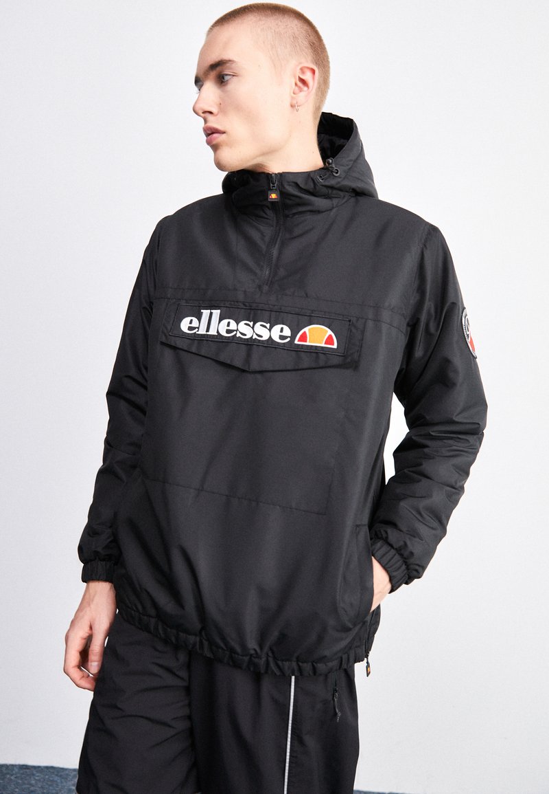 Giacca a vento nera con zip a metà, polsini elastici, tasca frontale e logo. Presenta un materiale resistente e un cappuccio regolabile.