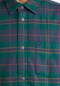 Kockás flaneling zöld, tengerészkék és narancssárga árnyalatokban, mellzsebbel és szürke gombos elülső záródással.