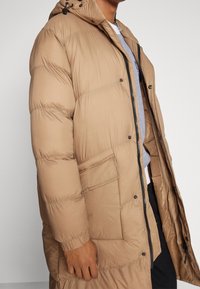 Beżowa kurtka puchowa z kapturem, przednim zamkiem błyskawicznym i guzikami. Posiada dużą kieszeń po lewej stronie oraz wzorzystą podszewkę.
