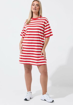 Donna in piedi, indossa un vestito a maglietta a righe orizzontali rosse e bianche e scarpe da ginnastica bianche, su uno sfondo chiaro e uniforme.