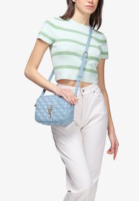 Borsa a tracolla trapuntata blu chiaro con bolle testurizzate e accessori in oro. Tracolla regolabile. Indossata da una persona con una maglietta a righe e pantaloni bianchi.
