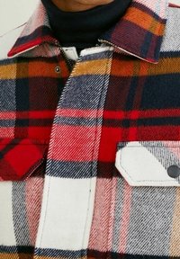 Rød, marineblå og hvidternet flannelskjorte med skjortekrave, to brystlommer og struktureret stof. Har en lynlås og trykknapper.