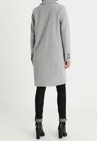 Lange grijze wollen jas met een kraag, geribde manchetten en een rechte snit. Draagt over zwart opgerolde jeans en zwarte laarzen met hak.