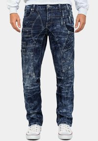 Pantaloni in denim blu scuro con più tasche, patch testurizzati e cuciture a contrasto. Design slim-fit con finitura usurata e leggera usura.
