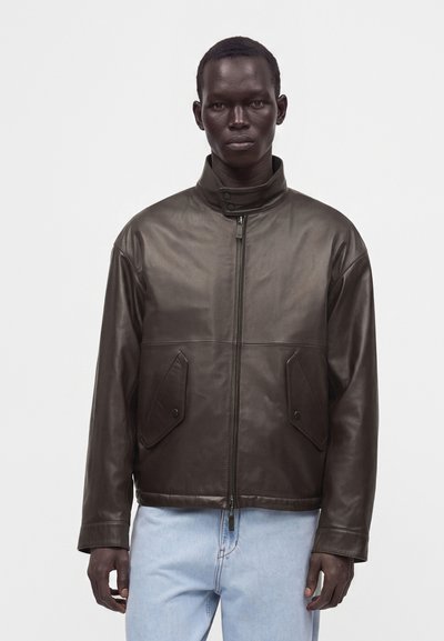 Mann trägt eine dunkelbraune Lederjacke mit hohem Kragen und Fronttaschen, kombiniert mit hellblauen Jeans, steht vor einem weißen Hintergrund.