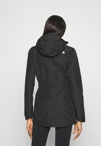 Personne aux longs cheveux portant une veste de pluie à capuche noire et des leggings noirs, vue de dos sur un fond blanc uni.