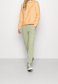 Persikkavärinen huppari, jossa on pieni sydänlogo, yhdistettynä vaaleanvihreisiin collegehousuihin, joissa on sivutaskut. Klassiset valkoiset ja vihreät tennarit täydentävät asun.