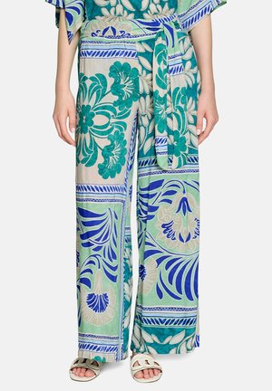 Weit geschnittene Hose mit grünen, blauen und beigen Blumen- und geometrischen Mustern, dazu ein passender Gürtel, kombiniert mit weißen Sandalen mit offenen Zehen.