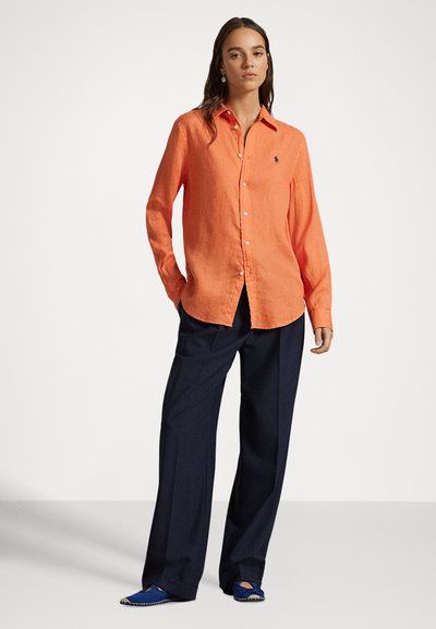 Sieviete stāv, tērpusies oranžā pogājamā kreklā, tumši zilās plaša piegriezuma biksēs un zilos slip-on apavos ar gaišu vienkāršu fonu.