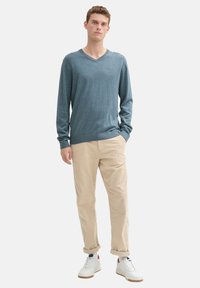 Maglione lunga manica a V di un blu teal chiaro, texture morbida, abbinato a chinos beige e sneakers bianche con accenti blu navy e rossi.