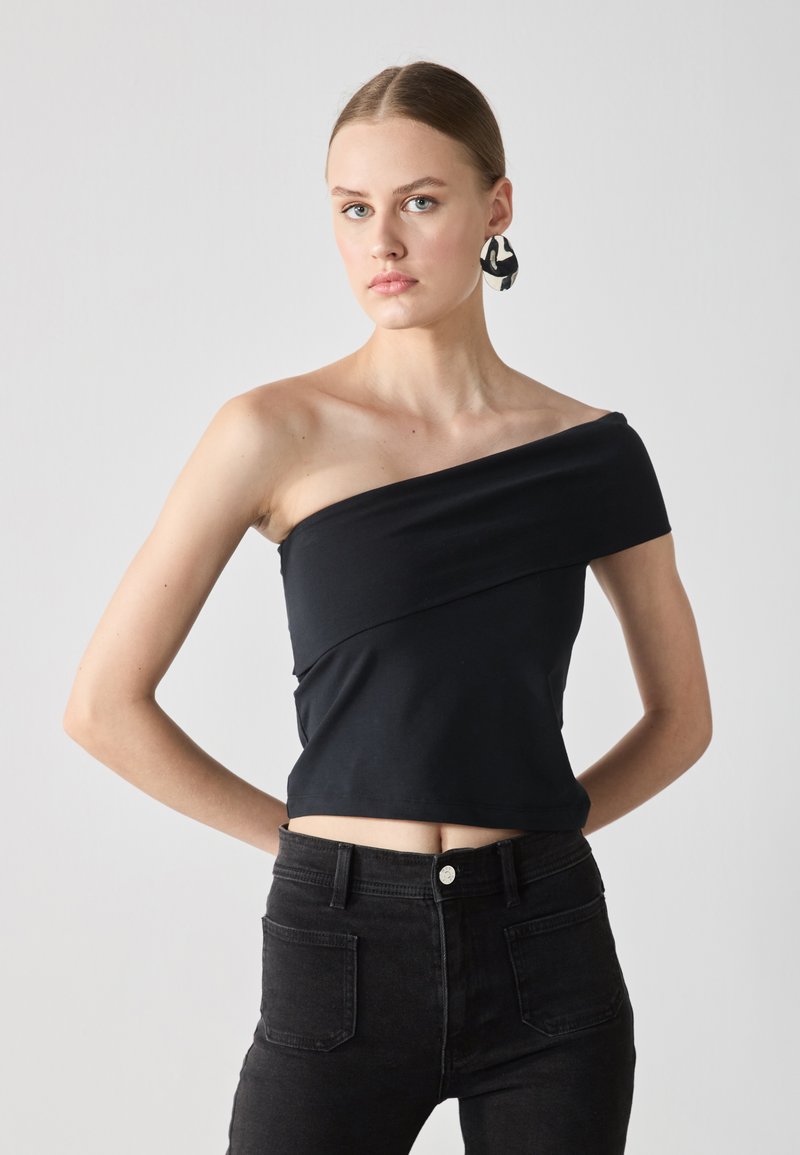 Černý crop top s odkrytými rameny vyrobený z hladkého materiálu, s diagonálním vzorem přes hruď. Kombinovaný s černými džíny s vysokým pasem.