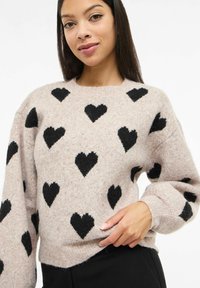 Mladá žena v béžovém svetru s černými srdíčky, stojící s jednou rukou držící lem svetru na bílém pozadí.