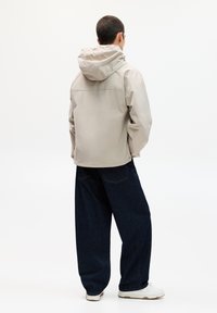 Veste imperméable beige avec capuche, au style ample. Portée avec un pantalon en denim bleu foncé à coupe décontractée et des baskets blanches.