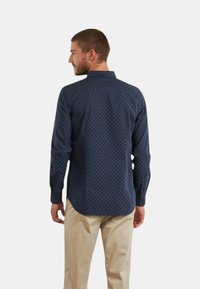 Camicia a maniche lunghe blu navy con piccoli pois bianchi, con colletto classico e orlo curvo, abbinata a pantaloni beige chiaro.