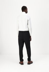 White long-sleeve shirt paired with black trousers, featuring a relaxed fit and side pockets, worn with brown leather shoes.