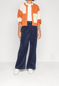 Oranžový a krémový pletený kardigan s předními knoflíky, spárovaný s námořnickými širokými manšestrovými kalhotami a bílými teniskami.