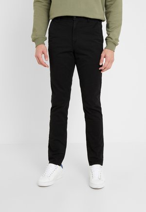 Mand iført sorte slim-fit bukser, hvide sneakers og en olivengrøn sweatshirt, stående på en ensfarvet lys baggrund.