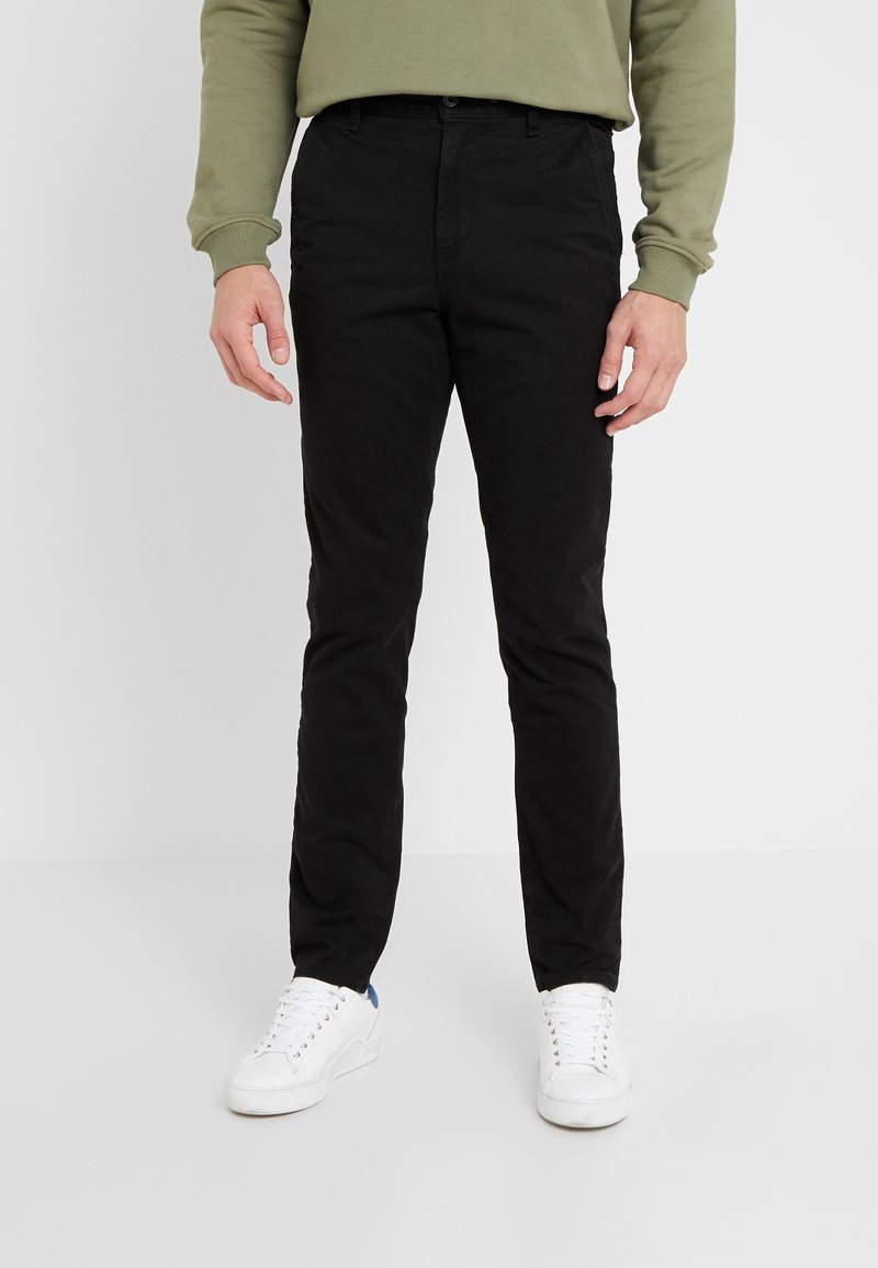 Homme portant un pantalon slim noir, des baskets blanches et un sweat-shirt vert olive, debout sur un fond clair uni.