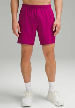 Mand iført magenta shorts, hvide crew sokker og hvide løbesko står på en neutral baggrund.