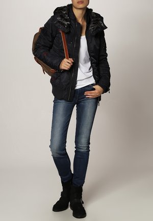 Personne portant une veste noire à capuche doublée de fourrure, une chemise blanche, un jean bleu, des bottes noires, et un sac à dos marron porté sur une épaule.