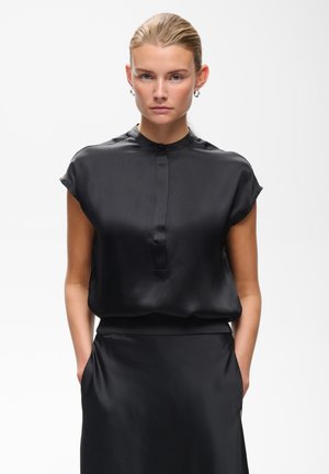 Femme aux cheveux plaqués en arrière portant un chemisier noir en satin à manches courtes et une jupe assortie, debout les mains dans les poches sur fond blanc.
