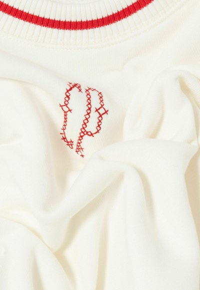 Vêtement en coton crème avec un col côtelé rouge. Présente un motif brodé rouge ressemblant à des lettres entrelacées sur le devant.