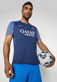 Mørkeblå sportstrøje med grå ærmer, der har "Qatar Airways" logo og rund halsudskæring. Spilleren holder en mønstret hvid og blå fodbold.