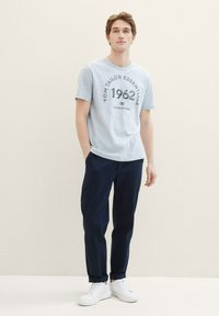 Svetlo modra majica s kratkimi rokavi in temnim tiskom "TOM TAILOR ESSENTIALS 1962" v kombinaciji s temno modrimi hlačami in belimi supergami.
