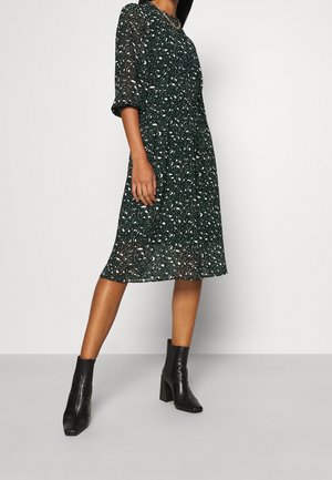Kvinde iført en mørkegrøn kjole i midilengde med et hvidt og beige abstrakt mønster og sorte ankelstøvler med hæl.