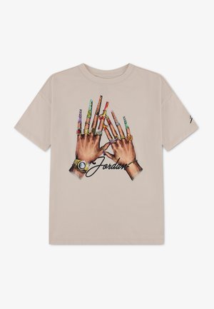 T-shirt beige avec un graphique de deux mains portant des bagues, des montres et de longs ongles décorés, ainsi que le mot "Jordan" en écriture manuscrite en dessous.