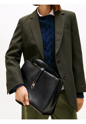 Personne portant un blazer olive à fines rayures et un pull en maille torsadée bleu marine, tenant un sac à main en cuir noir avec une fermeture dorée.