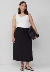 Zwarte, knielange rok met een elastische tailleband, gecombineerd met een witte tanktop. Houdt een gestructureerde zwarte accessoire vast. Platte sandalen.