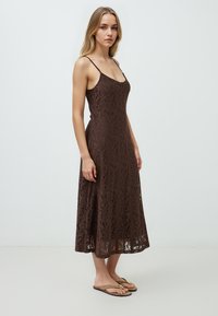 Abito midi marrone in pizzo con spalline sottili, caratterizzato da un motivo floreale e una silhouette fluida. Indossato con semplici ciabatte.