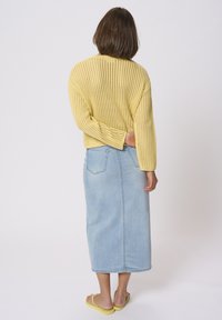 Suéter amarillo de punto con tejido abierto y mangas largas, combinado con una falda vaquera maxi de color azul claro. La modelo lleva chanclas amarillas.