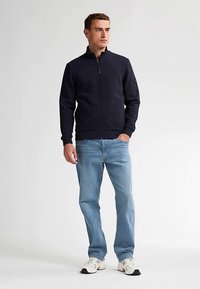 Mörkblå zip-up-jacka i mjukt tyg med ståkrage och ribbade muddar. Kombinerad med ljusblå jeans och vita sneakers.