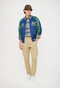 Modra satinasta bomber jakna z zelenimi rokavi, vezeninami, karirasto srajco, bež cargo hlačami in belimi športnimi copati. Rjav klobuk zaokroži obleko.