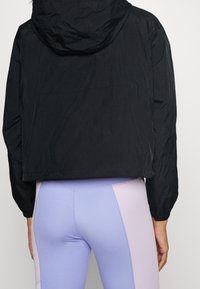 Svart kort jacka med texturerad yta och huva, tillsammans med ljuslila och rosa leggings med ett geometriskt färgblockdesign.