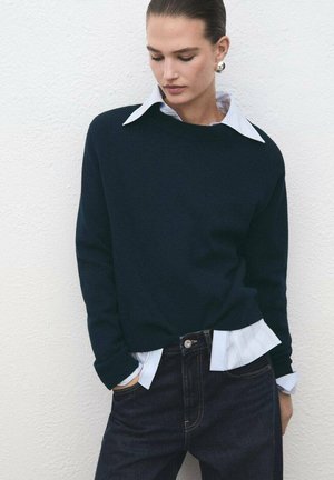 Maglione blu navy con scollo rotondo indossato sopra una camicia bianca con colletto. La camicia presenta delle righe blu. Il modello indossa jeans scuri con un bottone.