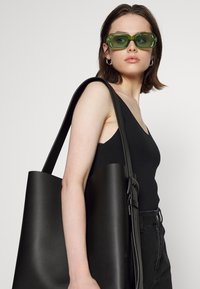 Bolsa tote preta em couro com textura suave, abertura ampla e alça longa para o ombro. O modelo usa óculos de sol verdes e uma camisola preta.