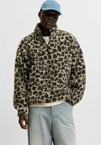 Pels-lignende leopard-print fleece-pullover med halvlynlås, høj krave og lange ærmer, sat sammen med lyseblå denimbukser og en blå kasket.