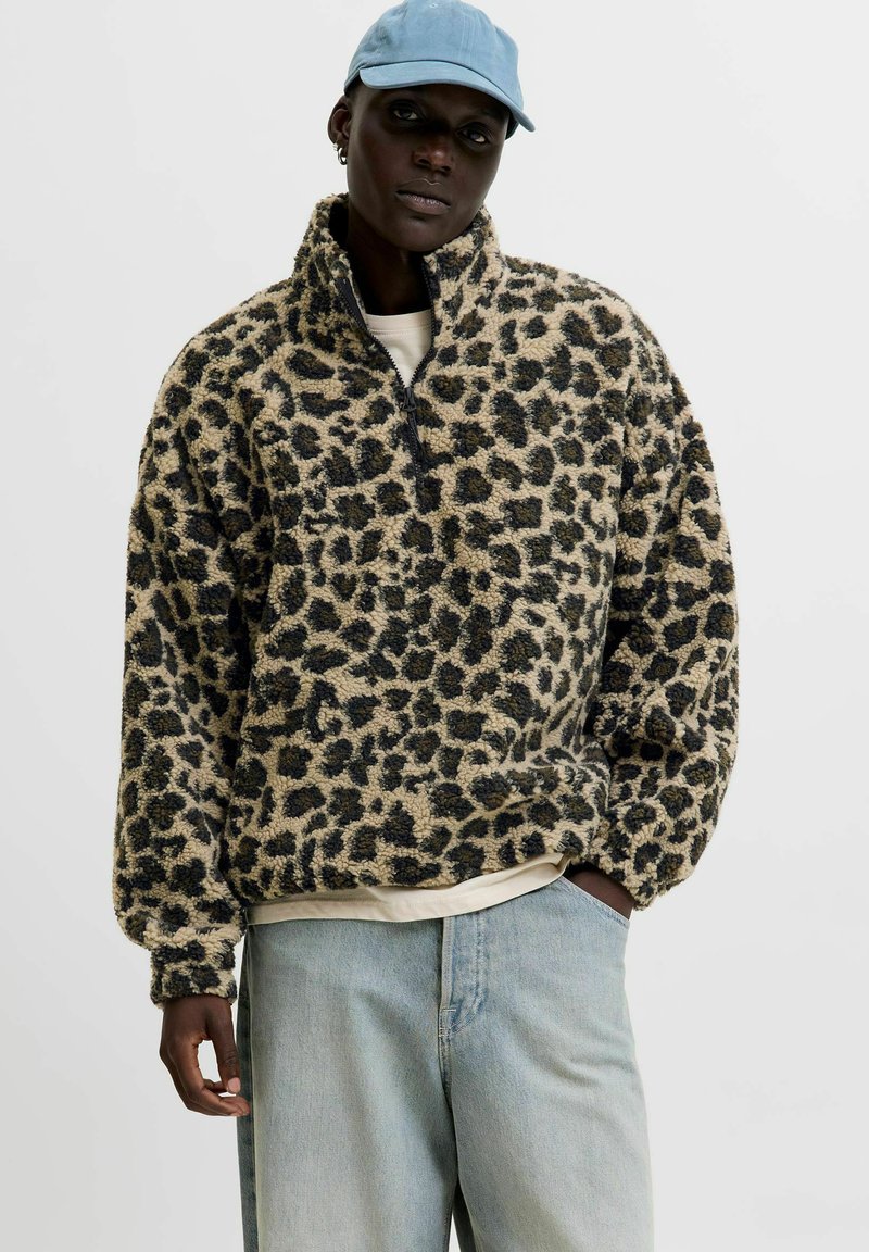 Pels-lignende leopard-print fleece-pullover med halvlynlås, høj krave og lange ærmer, sat sammen med lyseblå denimbukser og en blå kasket.
