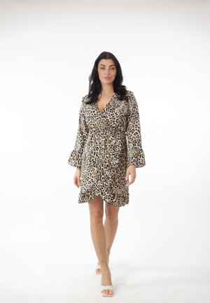 Kvinde med mørkt hår iført en slå-om-kjole med leopardprint og hvide åbne hæle, stående mod en ensfarvet hvid baggrund.