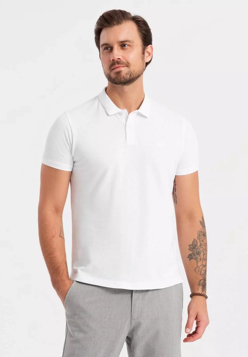 Vit polo-shirt med korta ärmar, platt krage och subtil textur. Kombinerad med ljusgrå byxor. Minimalistisk design, inga synliga logotyper.
