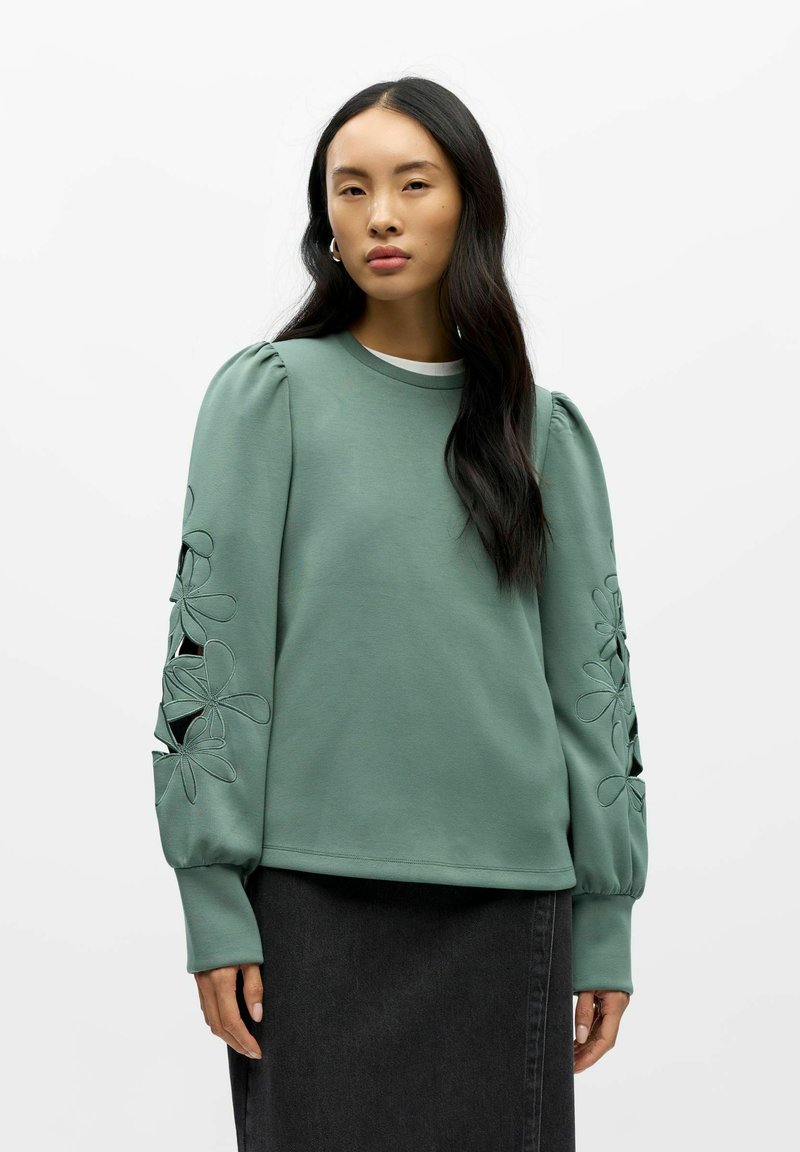Sweat-shirt vert avec des manches bouffantes, présentant des découpes florales et des détails brodés. Associé à une jupe gris foncé.
